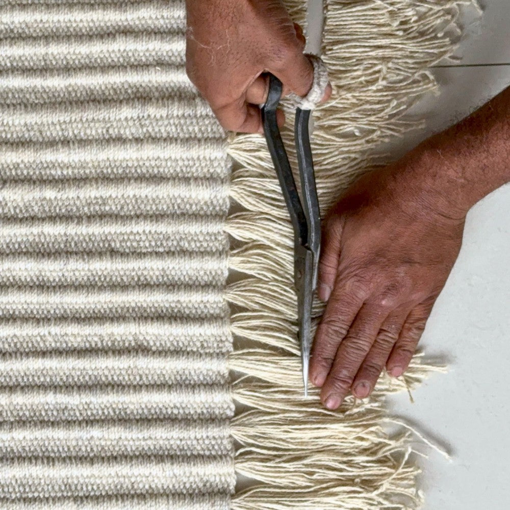 Artisan hand-finishing the fringe and binding of a flatweave daabi rug in ivory by House of Bohi, completing the final details of the handwoven piece.