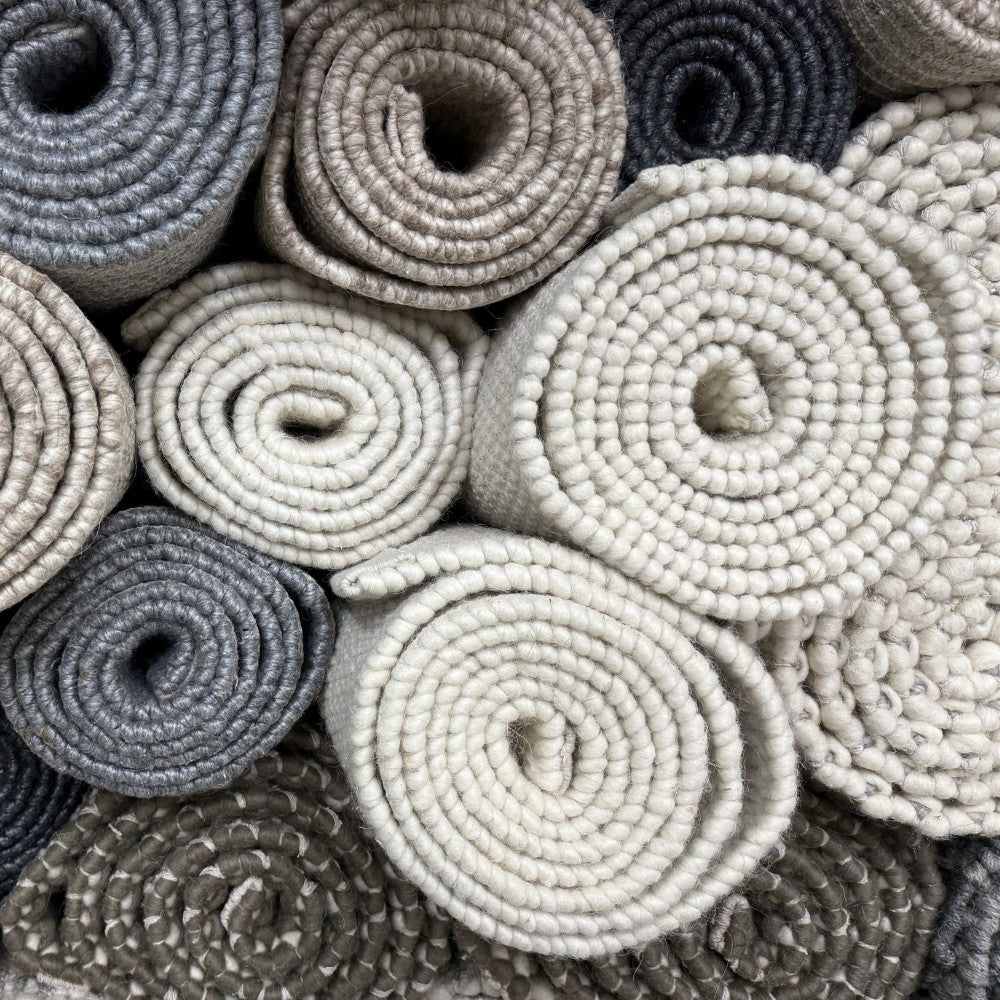 Stack of hand-woven flatweave rugs by House of Bohi, rolled and displayed in natural tones of ivory, stone, charcoal and grey wool and jute.