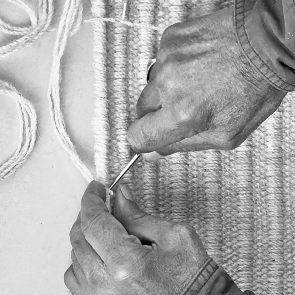 Black and white photograph of a master artisan edge binding the daabi flatweave wool rug at the House of Bohi workshop in Bhadohi, India, showing the generational craftsmanship behind every piece.