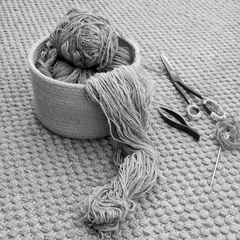 Black and white still-life of traditional rug-making tools for House of Bohi, showing a woven basket of wool yarn alongside needles, tweezers and scissors resting on a handwoven rug.
