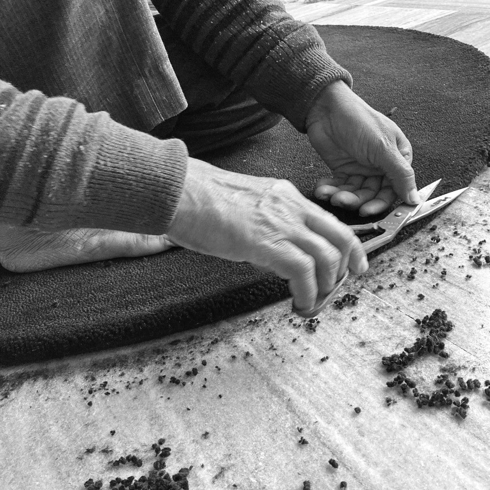 Black and white photograph of an artisan hand-finishing a wool rug with scissors for House of Bohi, carefully trimming the pile to achieve the refined quality behind every handmade piece.