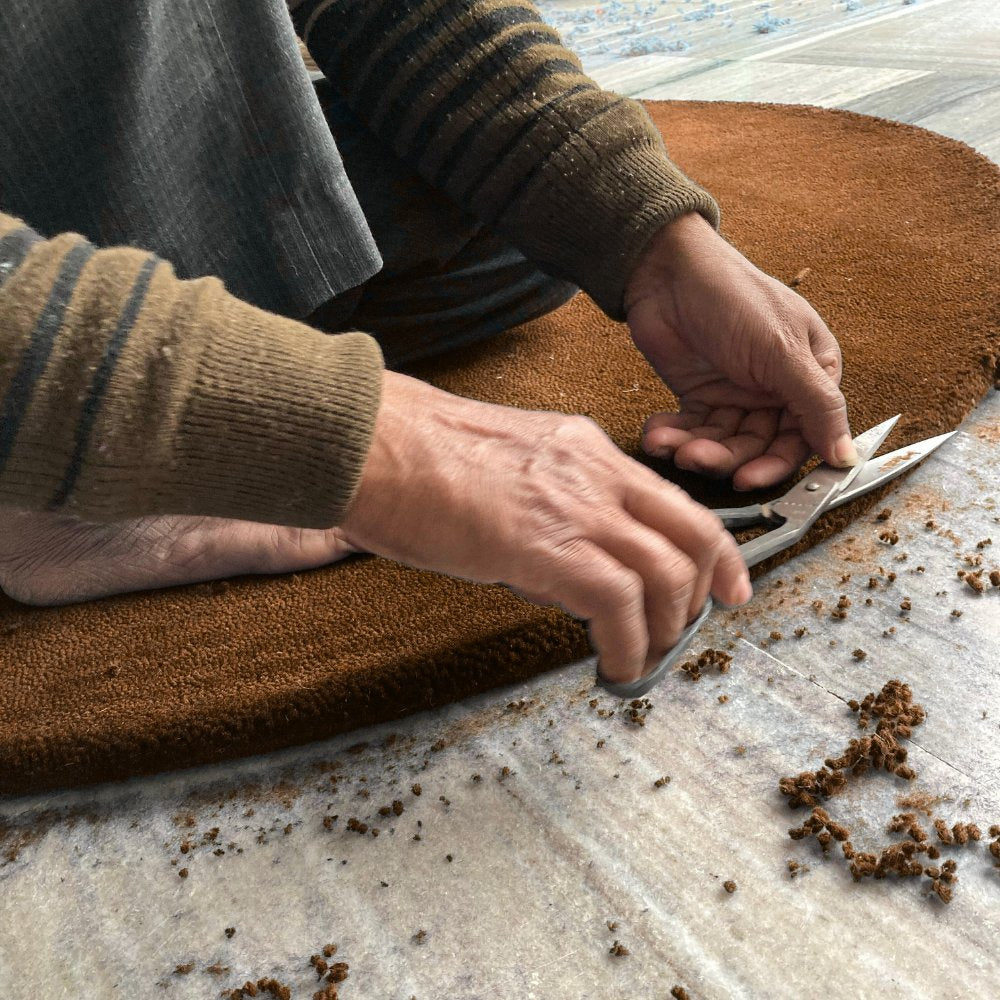 Artisan hand-finishing the edge of a custom round rug in brown by House of Bohi, trimming the wool pile with scissors to achieve a precise shape and size.