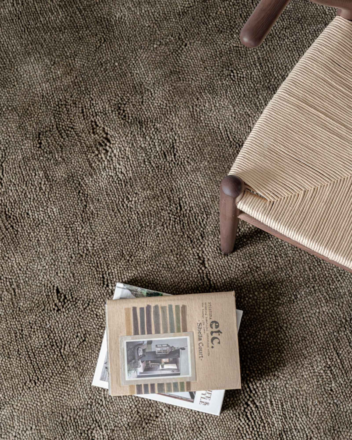 Overhead styled view of Raya Olive wool rug by House of Bohi, featuring a thick, nubby handloomed pile in muted olive green, beside a woven natural-fibre chair and stacked design magazines.