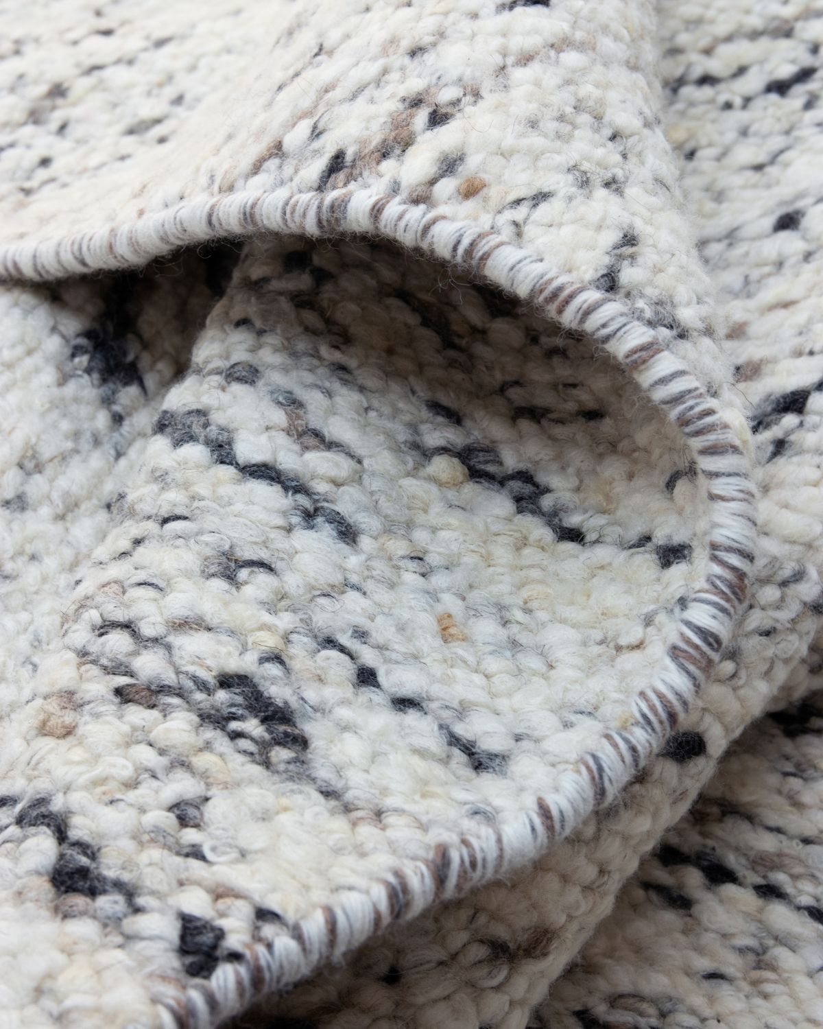Close-up of the hand-finished bound edge of Taalo Ashstone by House of Bohi, showing the rich texture of its hand-carded wool flatweave in natural undyed ivory, ash grey, charcoal and earthy brown tones.