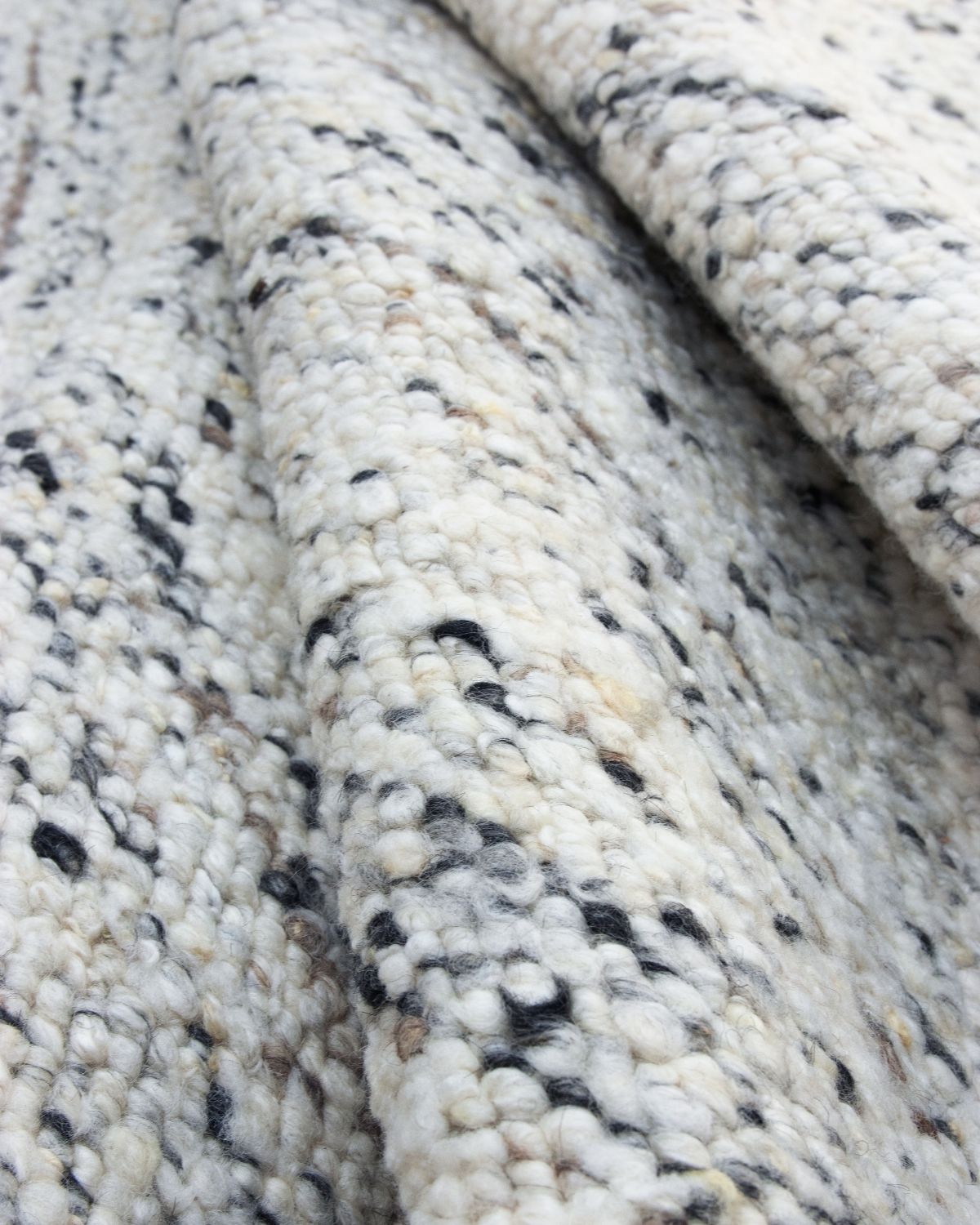 Folded close-up of Taalo Rug Ashstone, a handwoven undyed wool rug showcasing its plush hand-carded wool texture, a plush flatweave construction and natural ivory, ash grey and charcoal flecks.