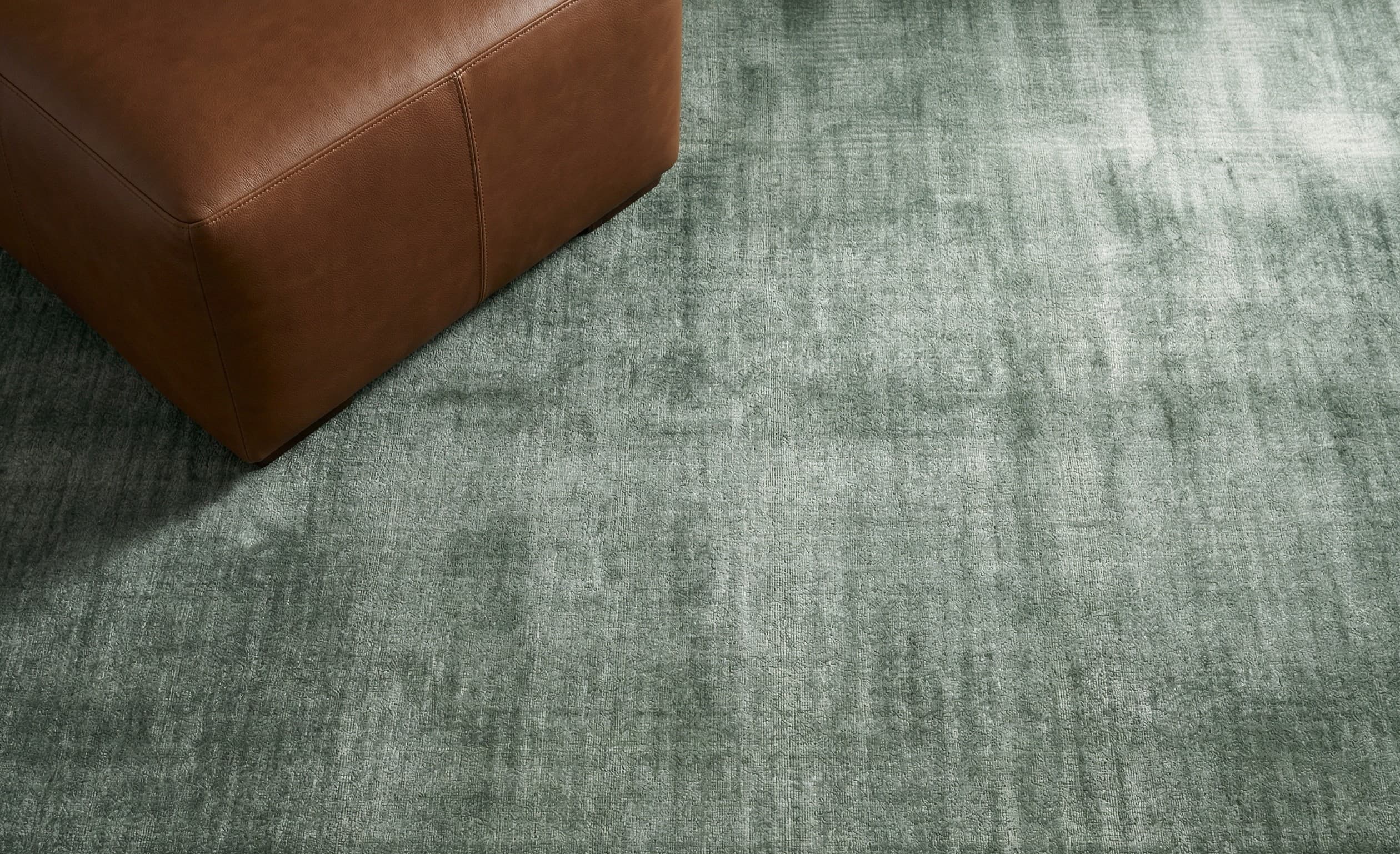Noor hand-loomed tencel rug in sage green by House of Bohi, with a subtle lustrous sheen and a tan leather ottoman resting on the corner.